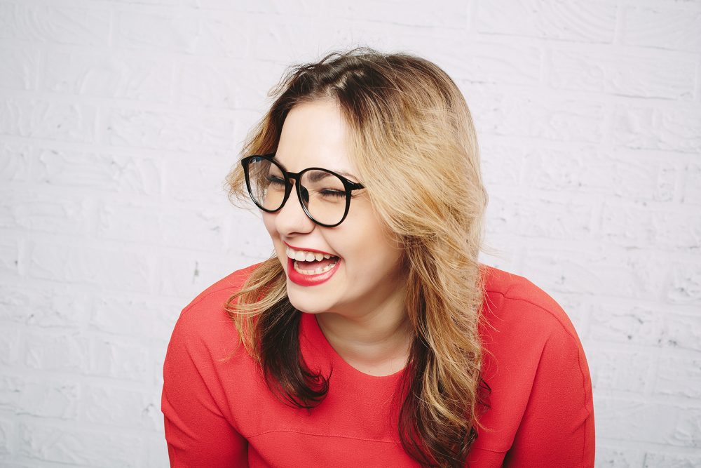 Laughing woman in red dress and glasses on white brick wall background